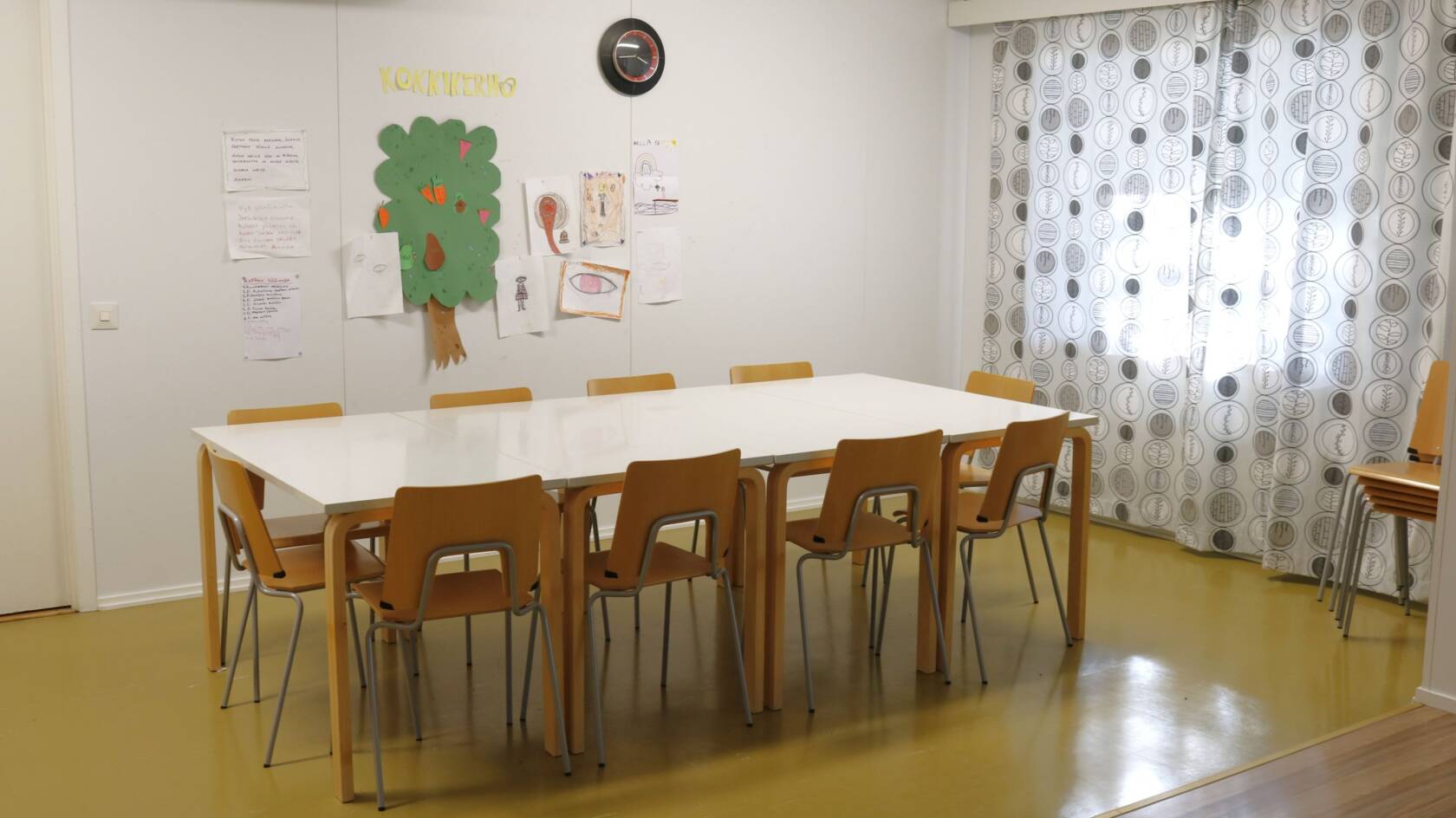 Iso pöytä ja tuoleja sen ympärillä. Vaalea seinä ja vaaleat verhot ikkunan edessä.