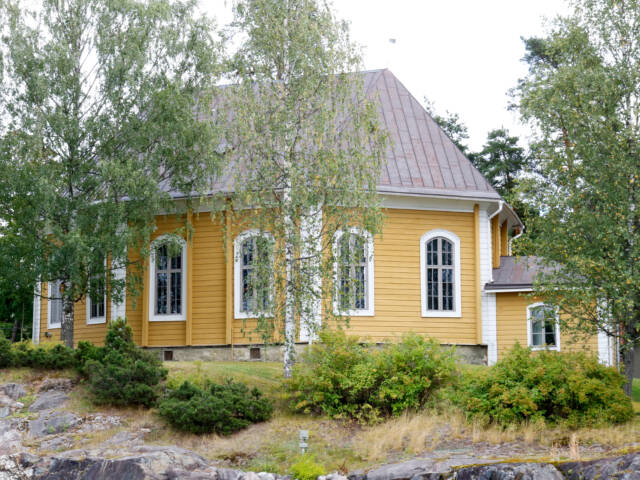 Heinolan kirkko on keltainen puukirkko. Ympärillä kasvaa koivuja ja pensaita.