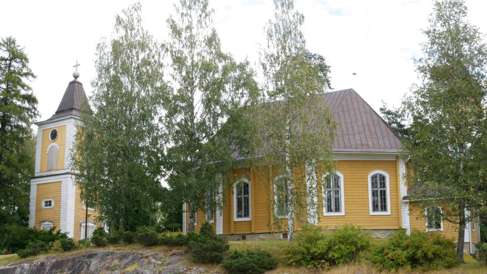Heinolan kirkko on keltainen puukirkko. Ympärillä kasvaa koivuja ja pensaita. Kuvassa on myös kellotapuli.