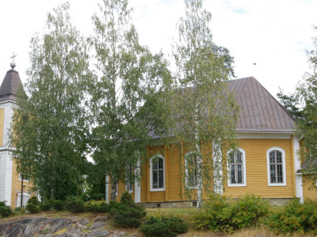 Heinolan kirkko on keltainen puukirkko. Ympärillä kasvaa koivuja ja pensaita. Kuvassa on myös kellotapuli.
