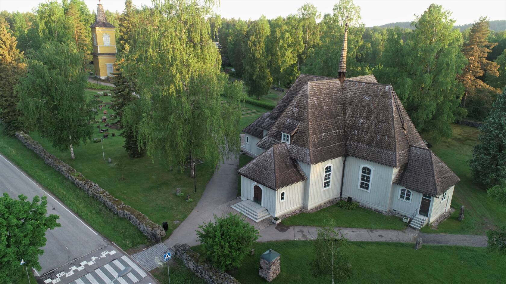 Heinolan pitäjänkirkko ja kellotapuli sekä Vanha hautausmaa ilmasta kuvattuna. Pitäjänkirkko on harmaa puurakenteinen ristikirkko, kahtamoinen.