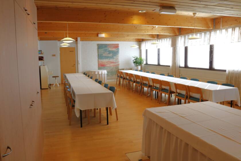 Pitkät pöydät ja tuoleja. Pöydillä valkoiset liinat. Takaseinällä värikäs ryijy ja sivuseinillä ikkunoita.