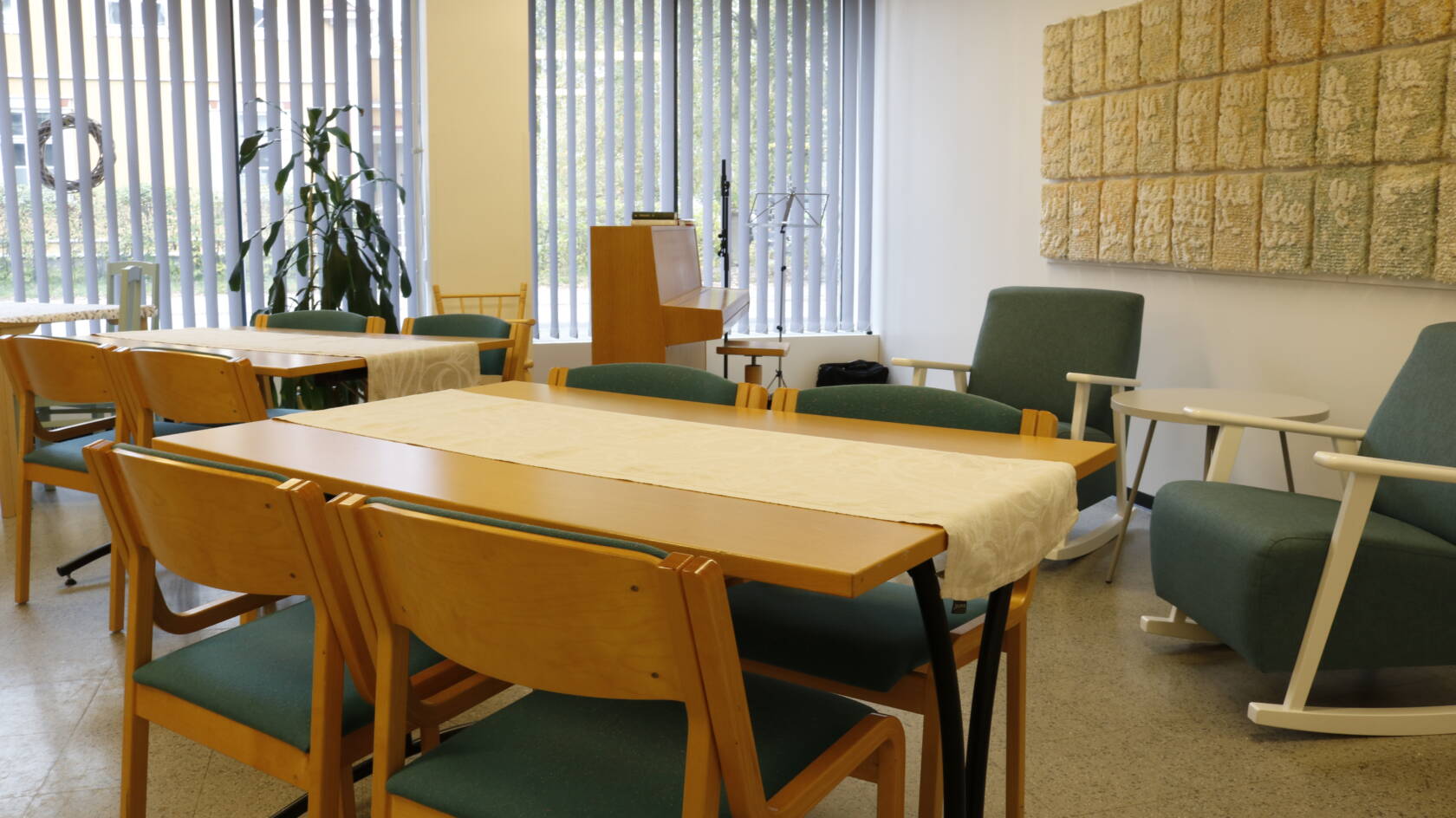 Pöytäryhmiä ja turkooseja pehmustettuja tuoleja sekä piano ikkunan ääressä.