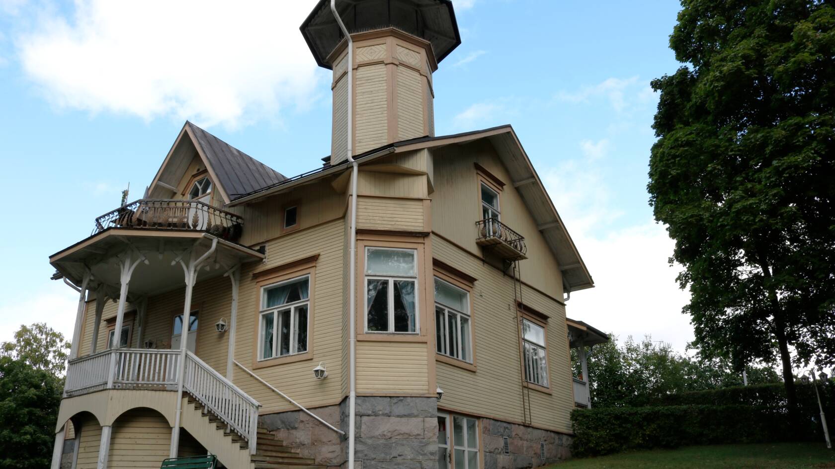 Vaaleanruskea kaksikerroksinen puuhuvila, jossa on torni, koristeelliset parvekkeet ja portaikko.