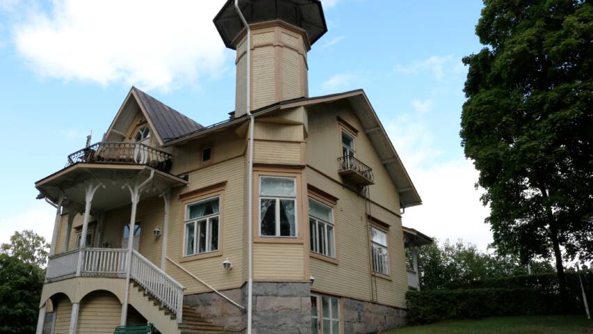Vaaleanruskea kaksikerroksinen puuhuvila, jossa on torni, koristeelliset parvekkeet ja portaikko.