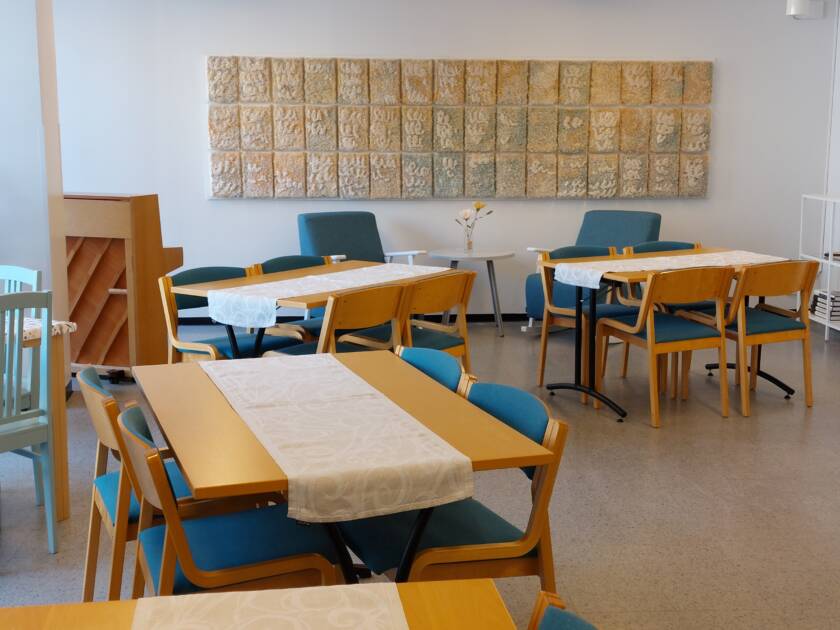 Pöytäryhmiä joissa turkoosit tuolit. Takaseinällä vaalea tekstiiliteos.