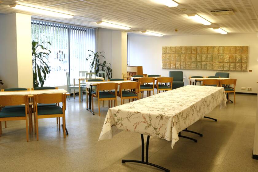 Pöytäryhmiä ja tuoleja sekä pitkä tarjoilupöytä. Takaseinällä suuri vaalea tekstiiliteos.