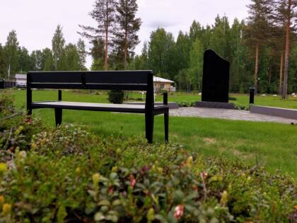 Puistonpenkki ja tumma muistomerkki, nurmikkoa ja kunttaa.
