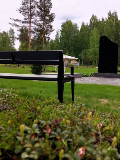 Puistonpenkki ja tumma muistomerkki, nurmikkoa ja kunttaa.