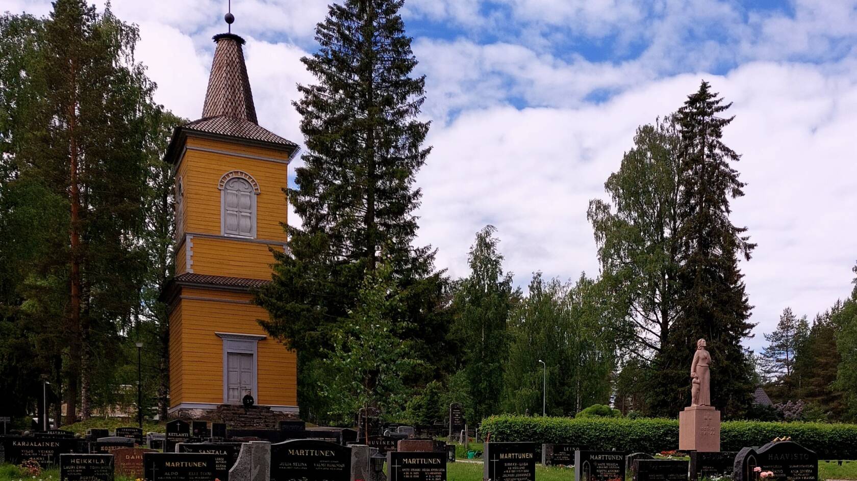 Hautakiviä sekä keltainen puinen kellotapuli ja puita Vanhalla hautausmaalla