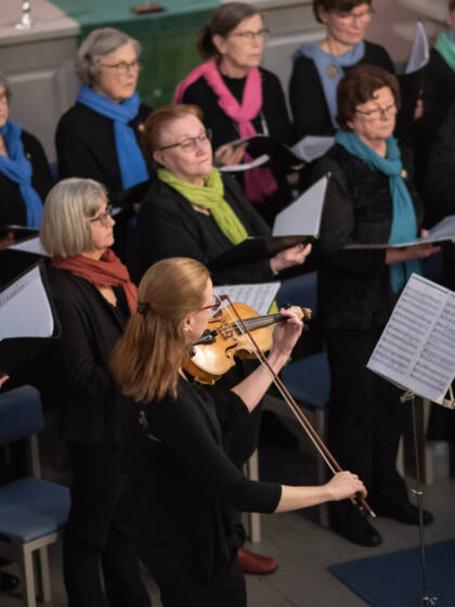 Kuorolaisia tummissa vaatteissa, värikkäät huivit kaulassa. Etualalla viulisti soittaa.