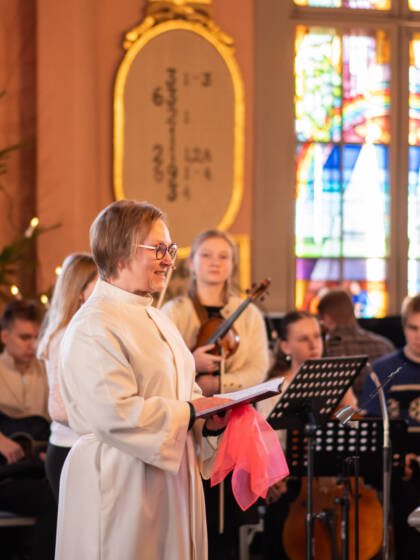 Liturgi kirkossa päällään valkoinen alba. Takana nuoria soittajia.