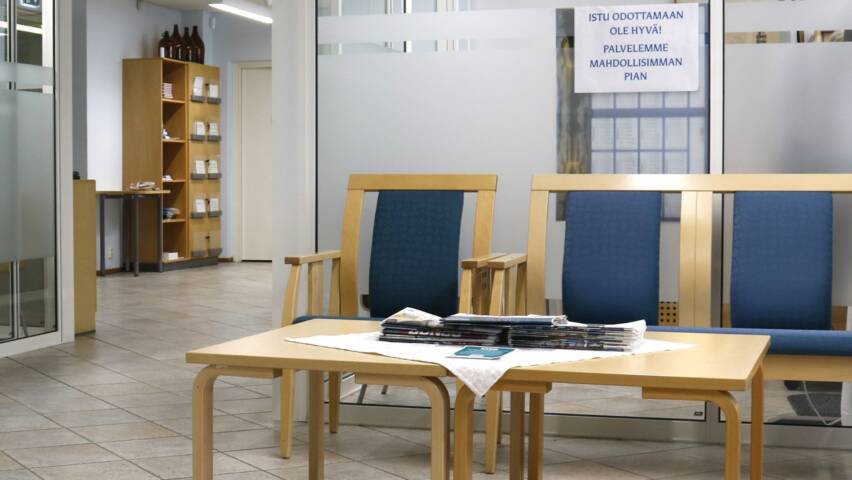 Aulatila, jossa on naulakko, sohva ja tuoli sekä pöytä. Lasiseinän takana vastaanottohuone.