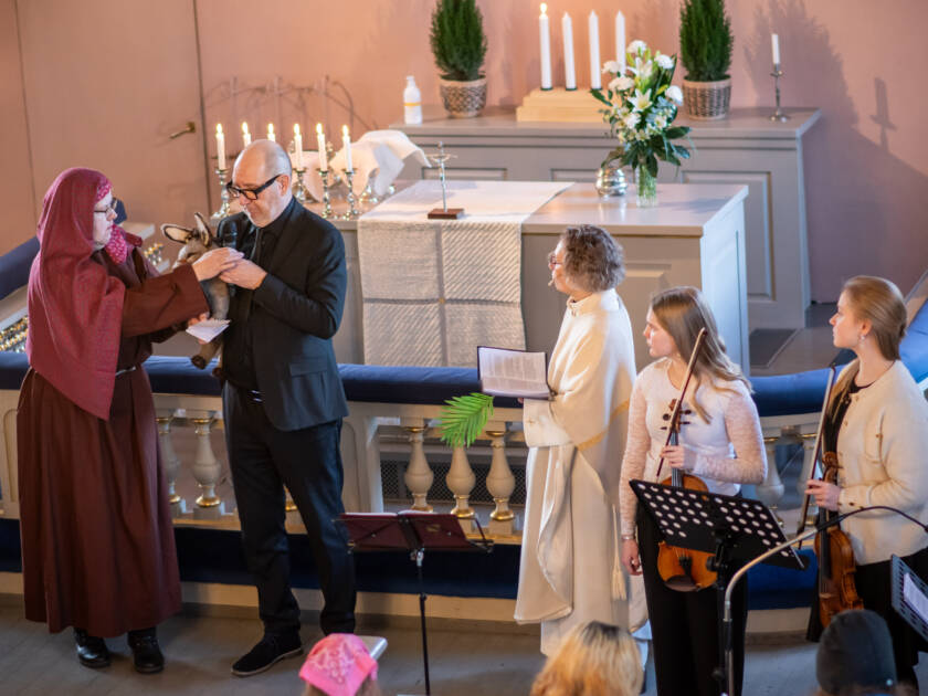 Liturgi ja kaksi viulistia katsovat kahta henkilöä, jotka esittävät Raamatun kertomusta. Taustalla kirkon alttari.