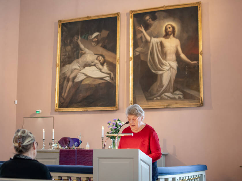 Henkilö kirkon etuosassa puhumassa, taustalla Kristusta esittävät kaksi alttarimaalausta. Penkissä istuu toinen henkilö.