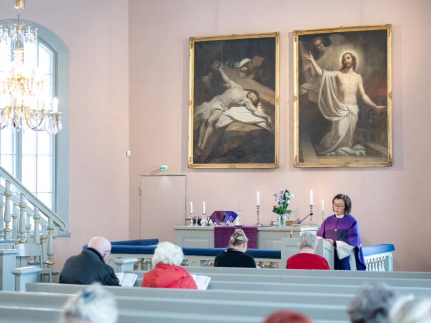 Ihmisiä istumassa kirkon penkeillä, pappi edessä puhumassa. Taustalla Kristusta ristillä ja ylösnousseena esittävät alttarimaalaukset.