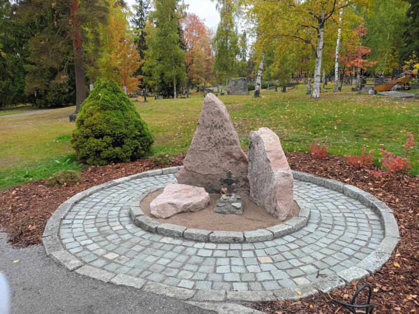 Muistomerkki hautausmaalla. Kolme suurta kivipaasia ja niiden keskellä metallinen risti. Ympärillä ympyränmuotoinen kiveys ja puistomaista hautausmaata.