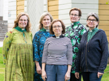 Viisi henkilöä seisoo rivissä ja katsoo kameraan, neljällä on vihreä diakonin kaulus ja yhdellä musta papin kaulus. Taustalla kesäinen kirkkopuisto ja kellotapulin seinää.