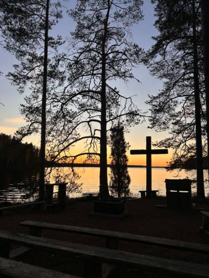 Leirikeskusken nuotiopaikka iltahämärällä, taustalla iso puinen risti ja järvi.