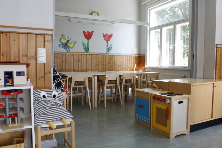 Lasten kerhotila, jossa on suuret ikkunat, pöytä ja tuoleja sekä leluja.