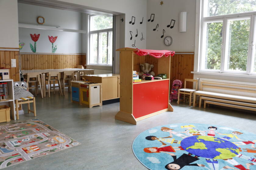 Lasten kerhotila, jossa on suuret ikkunat, pöytä ja tuoleja, leikkikioski ja värikkäitä mattoja lattialla.